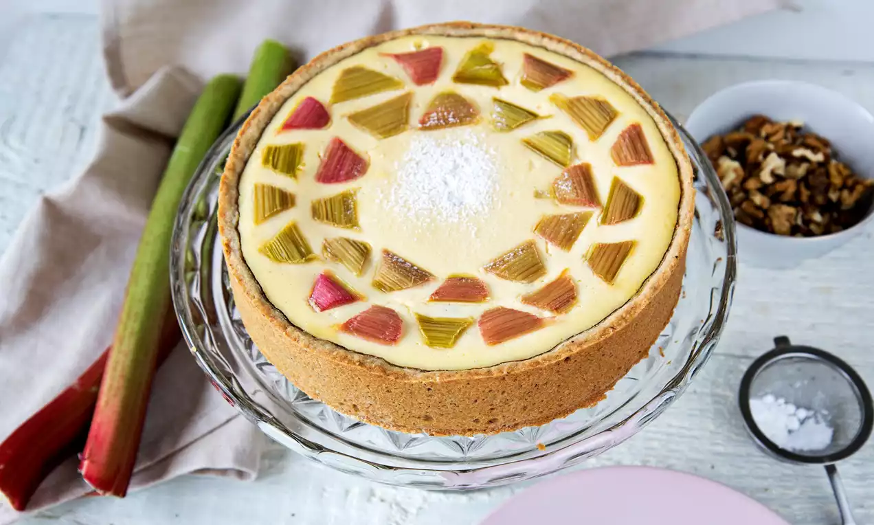 Ricottový koláč na skleněném talíři, zlatohnědý okraj, světlý krém posetý růžovými a zelenými rebarborovými kousky, doplněný vlašskými ořechy