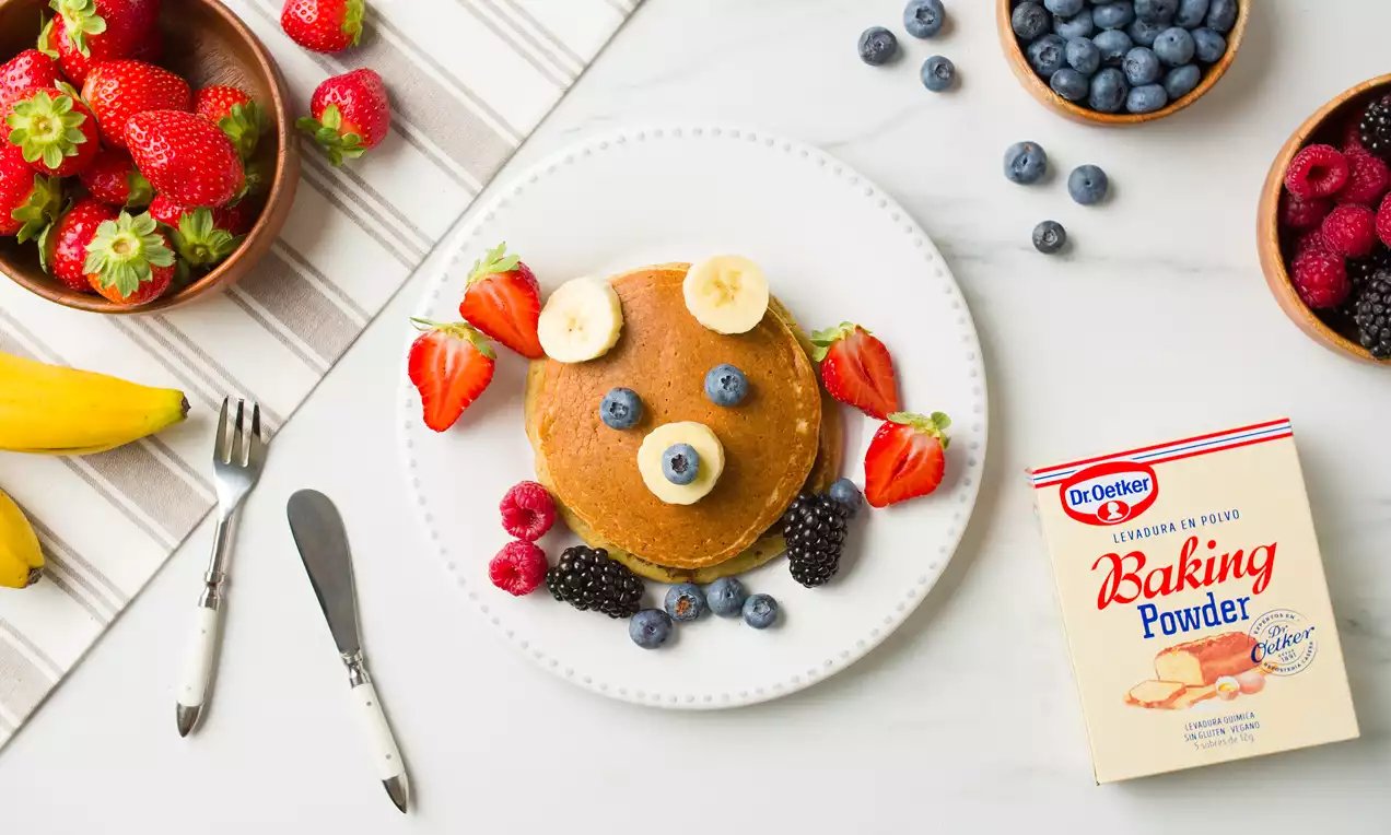 Tortitas circulares doradas decoradas con rodajas de plátano, fresas, moras y arándanos sobre un plato blanco en una mesa con frutas