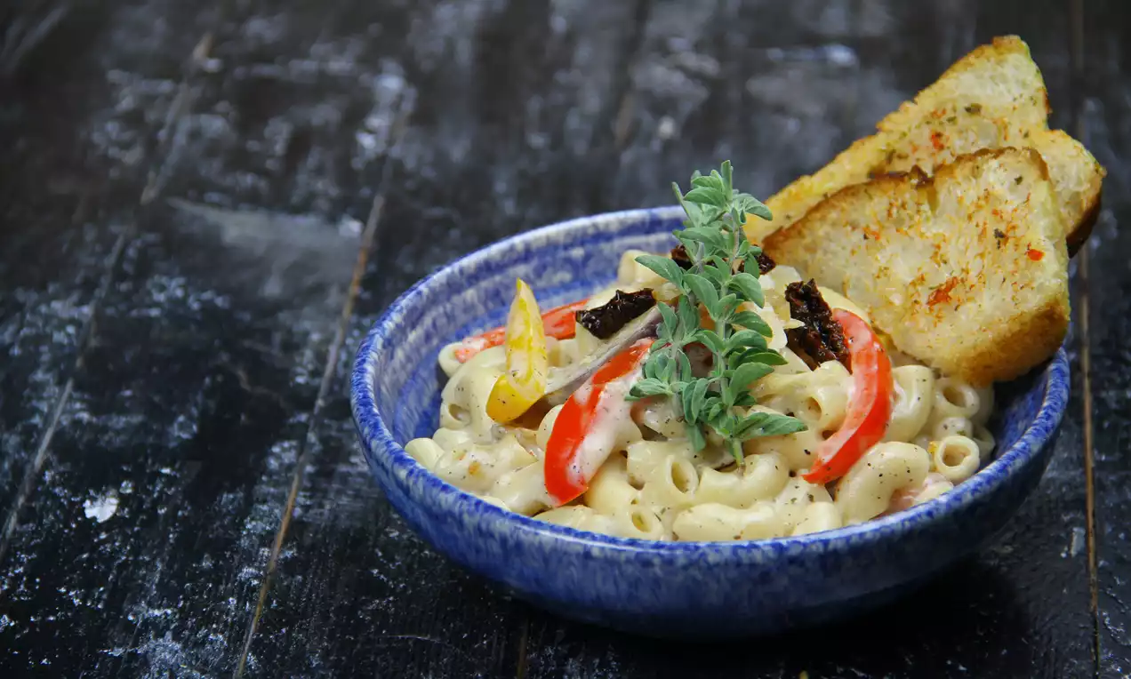 A bowl of creamy pasta with curved grilled red and yellow vegetable strips, garnished with fresh herbs and served with golden toasted bread