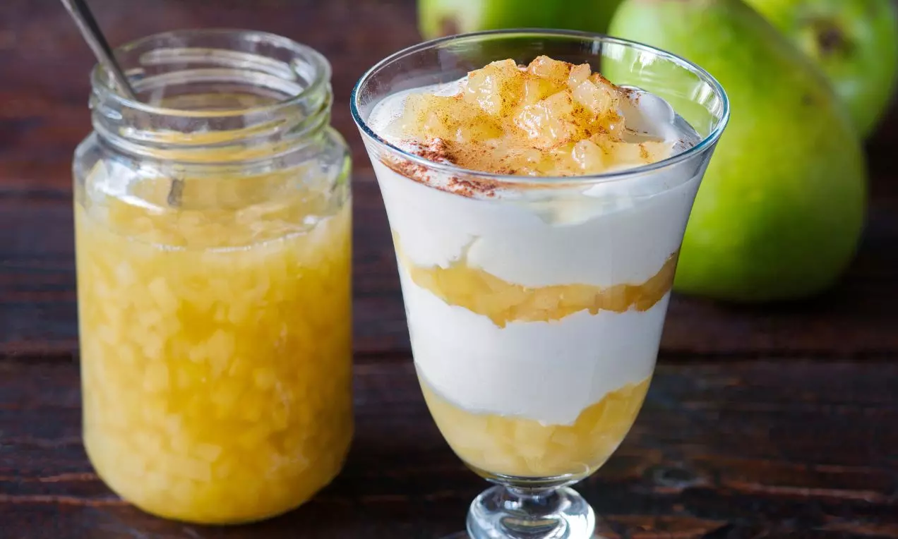 Vysoký sklený pohár s vrstvami bieleho krému a zlatistej hruškovej zmesi, ozdobený kúskami hrušky a škoricou, vedľa zaváraninového pohára a zelených hrušiek
