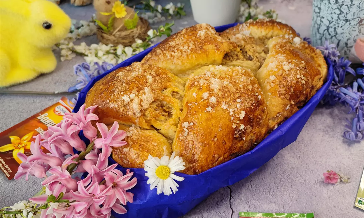 Златисто-кафяв козунак с плетка и орехова плънка, поръсен с кристална захар, поставен в синя хартиена основа с пролетни цветя около него