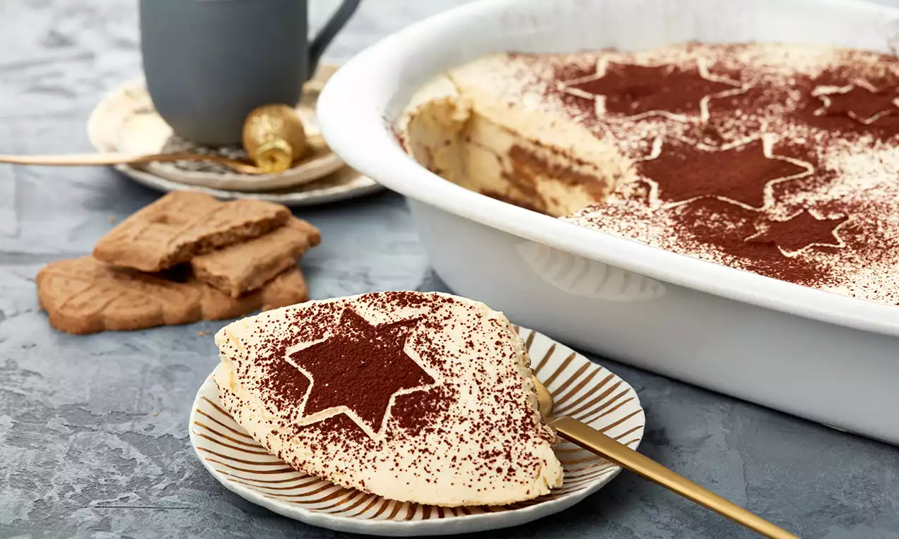 Ein Stück Tiramisu mit heller Creme und spekulatiusgewürztem Boden, bestäubt mit Kakaopulver und Sternmotiv auf einem Teller mit goldener Gabel