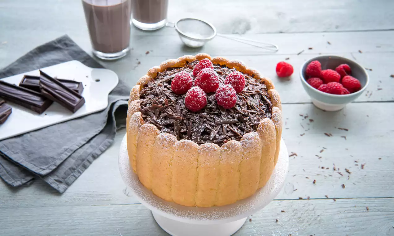 Ronde charlottetaart omringd door lichtbruine lange vingers, gevuld met chocoladeschaafsel en rode frambozen, op een wit taartbord