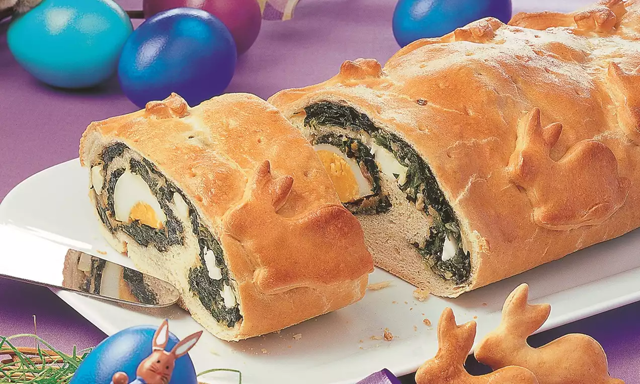 Ruladă aurie cu aluat pufos, umplutură de spanac verde intens și ouă fierte, așezată pe un platou alb lângă ouă colorate și decorațiuni de Paște