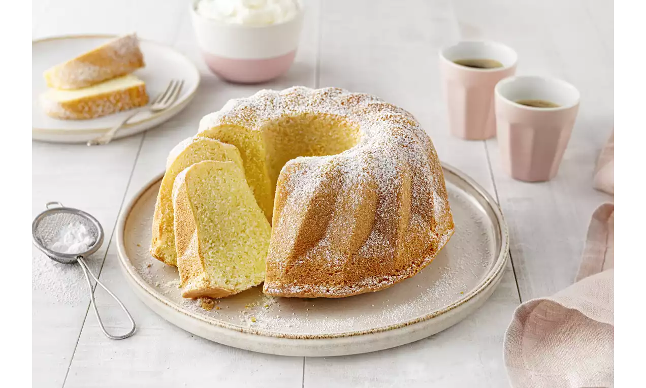 Sandkuchen Gugelhupf mit goldbrauner Kruste und leichter Puderzuckerschicht, in Scheiben auf weißem Teller neben roten Beeren angerichtet