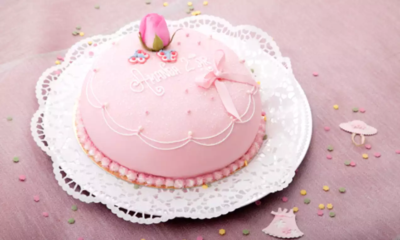 Rund prinsesstårta täckt med ljusrosa marsipan, dekorerad med en marsipanros, pärlband, rosett och text på en vit spetsduk
