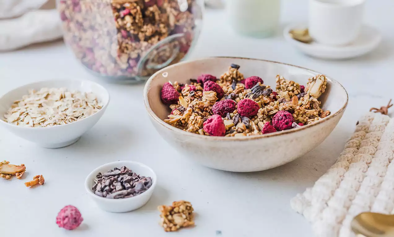 Granola zlatavě hnědá s hrudkami různých velikostí, mezi nimi sušené červené maliny, podávaná v béžové keramické misce na světlém stole