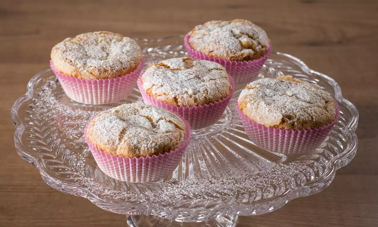 Muffin dorati e soffici spolverati di zucchero a velo, avvolti in pirottini rosa su un'alzata di vetro trasparente lavorato