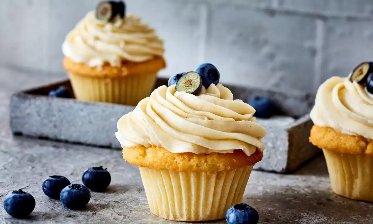 Cupcake doré avec glaçage vanillé ondulé, garni de myrtilles fraîches sur une table en pierre grise et un plateau en arrière-plan