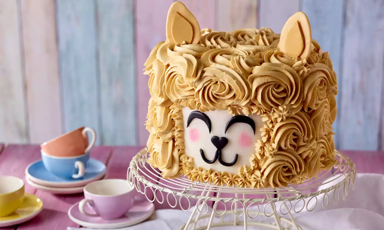 Runde Torte mit goldbrauner Buttercreme in lockigen Rosetten, einem weißen Gesicht mit Alpaka-Mimik und Ohren auf Metalltortenständer