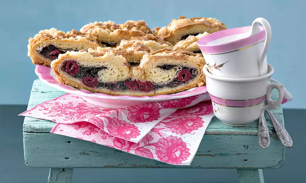 Plátky závinů se zlatavou kůrkou, tmavou makovou náplní a šťavnatými višněmi na růžovém talíři s květinovým ubrouskem na modrém stole