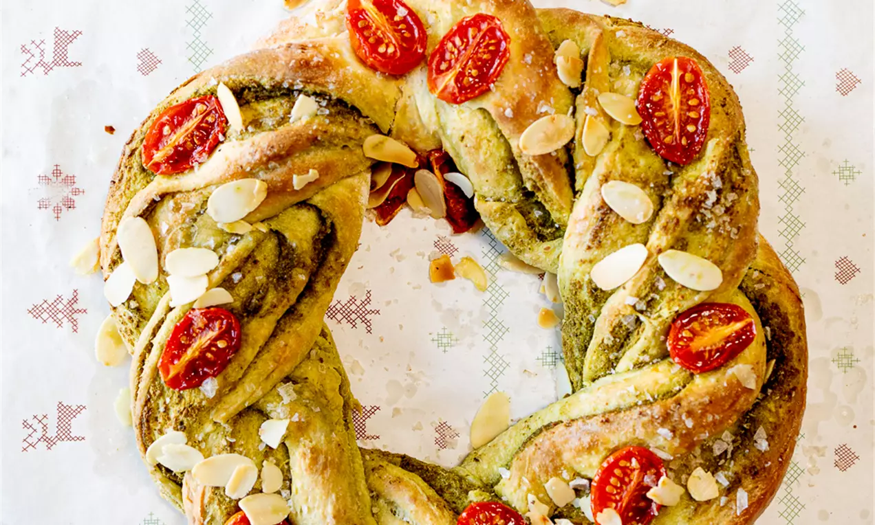 Pleteni kruh zlatnosmeđe boje s trakama zelene pesto kreme, dekoriran polovicama crvenih cherry rajčica i listićima badema na papiru s blagdanskim uzorkom
