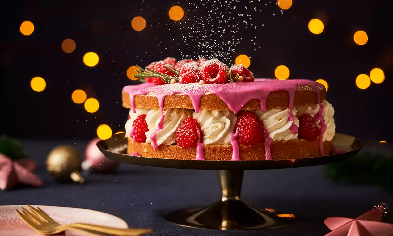 Kersttaart met luchtige lagen, roze glazuur, slagroomtoefjes, verse frambozen en dennentakjes op een goudkleurige taartstandaard