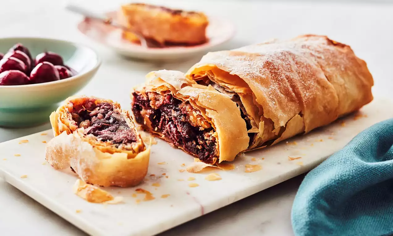 Golden-brown strudel with flaky pastry, filled with deep red cherries, chocolate, and marzipan, sliced on a marble board near a teal napkin