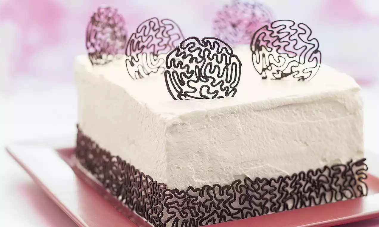 Dikdörtgen beyaz kremalı pasta, kenarları ve üstü zarif kıvrımlı siyah çikolata desenleriyle süslenmiş, kırmızı tabakta sunulmuş