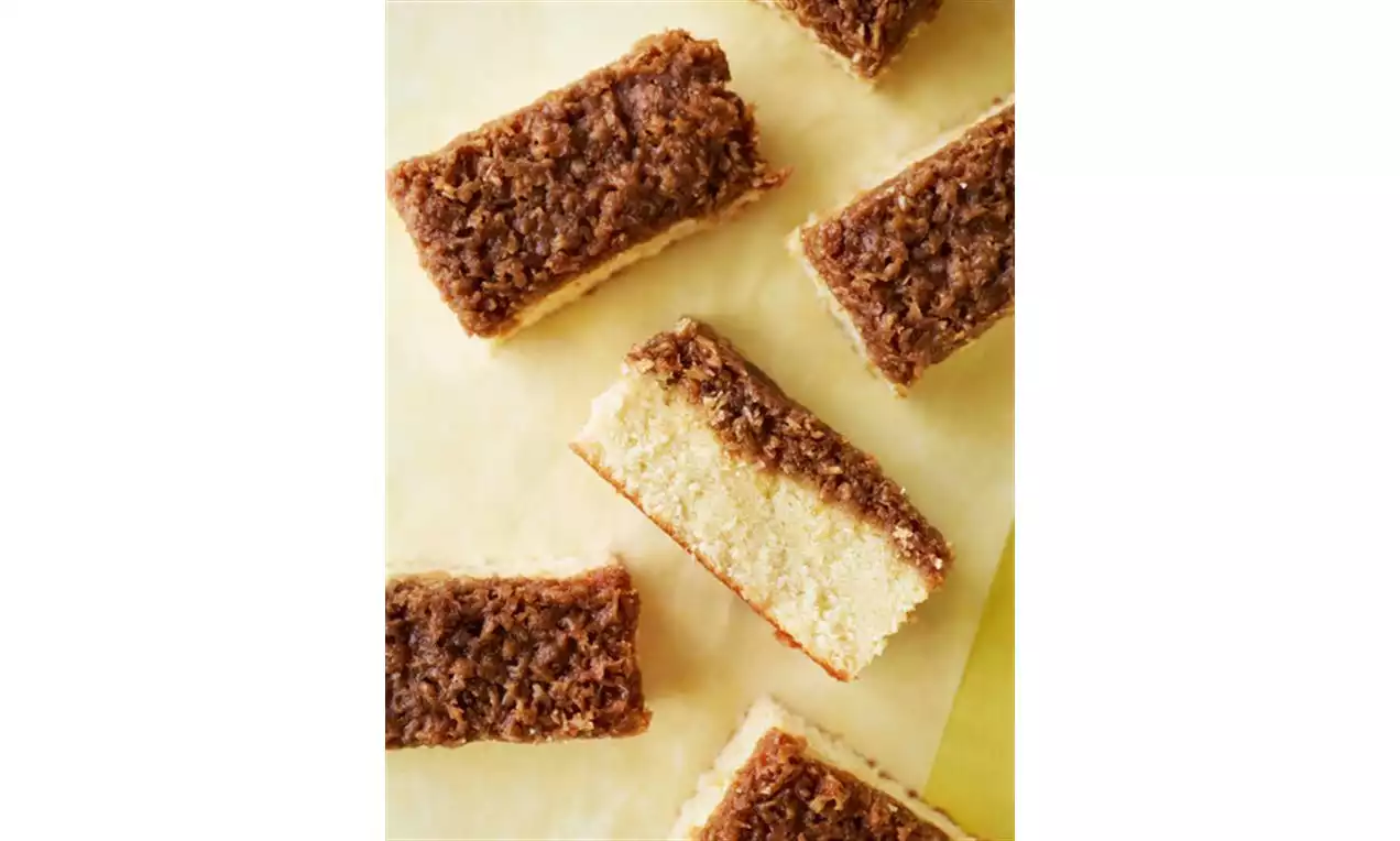 Rektangulære stykker med lys kakebunn og mørk brun kokosglasur ligger jevnt fordelt på lysegult bakepapir