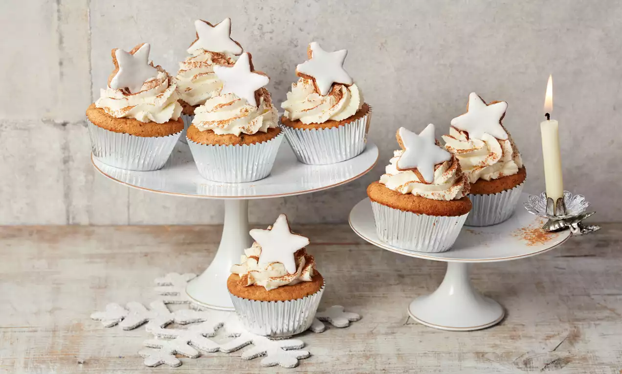 Goldbraune Weihnachtsmuffins mit weißer Cremehaube, bestäubtem Zimt und weiß glasierten Sternen auf weißen Tortenständern mit Kerze