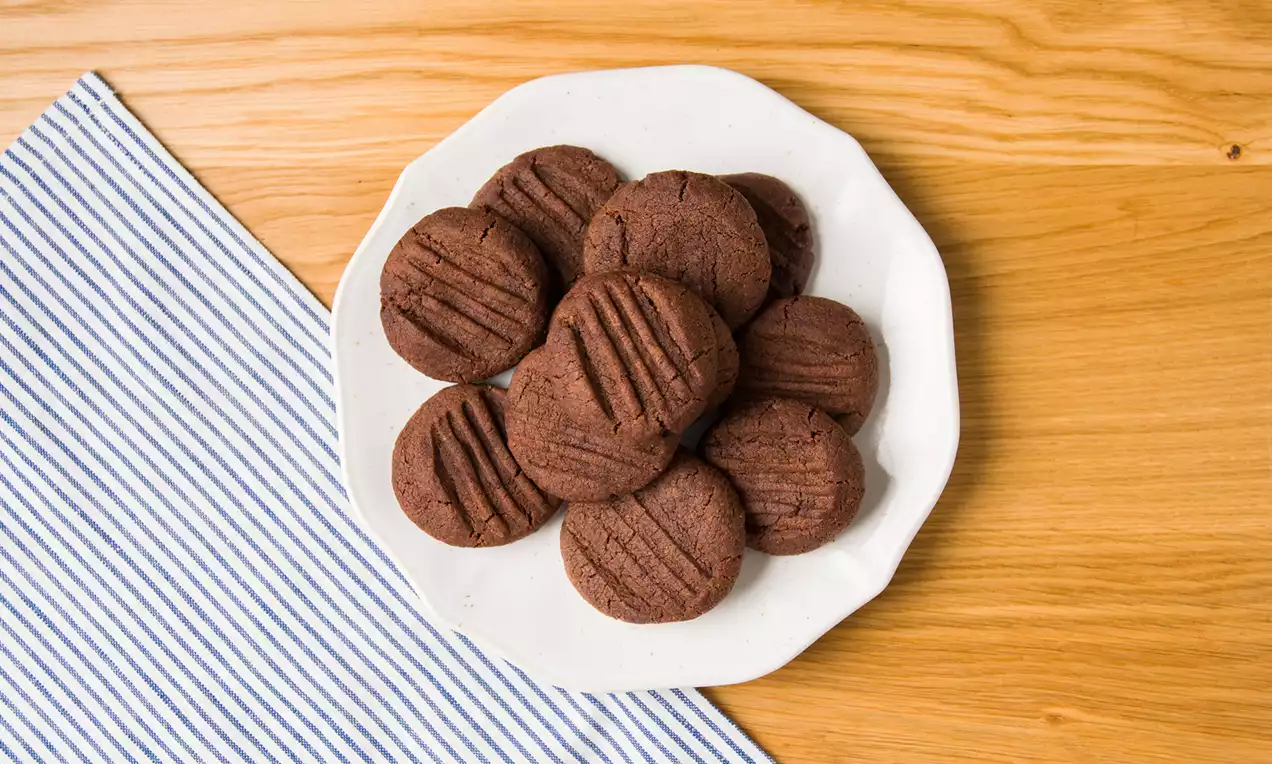 Galletas redondas de chocolate oscuro con textura estriada en su superficie, dispuestas sobre un plato blanco sobre una mesa de madera clara