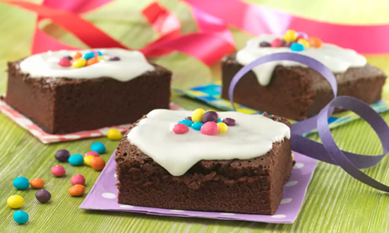 Brownies de chocolate oscuro con glaseado blanco brillante decorados con pequeños caramelos coloridos sobre bases de colores