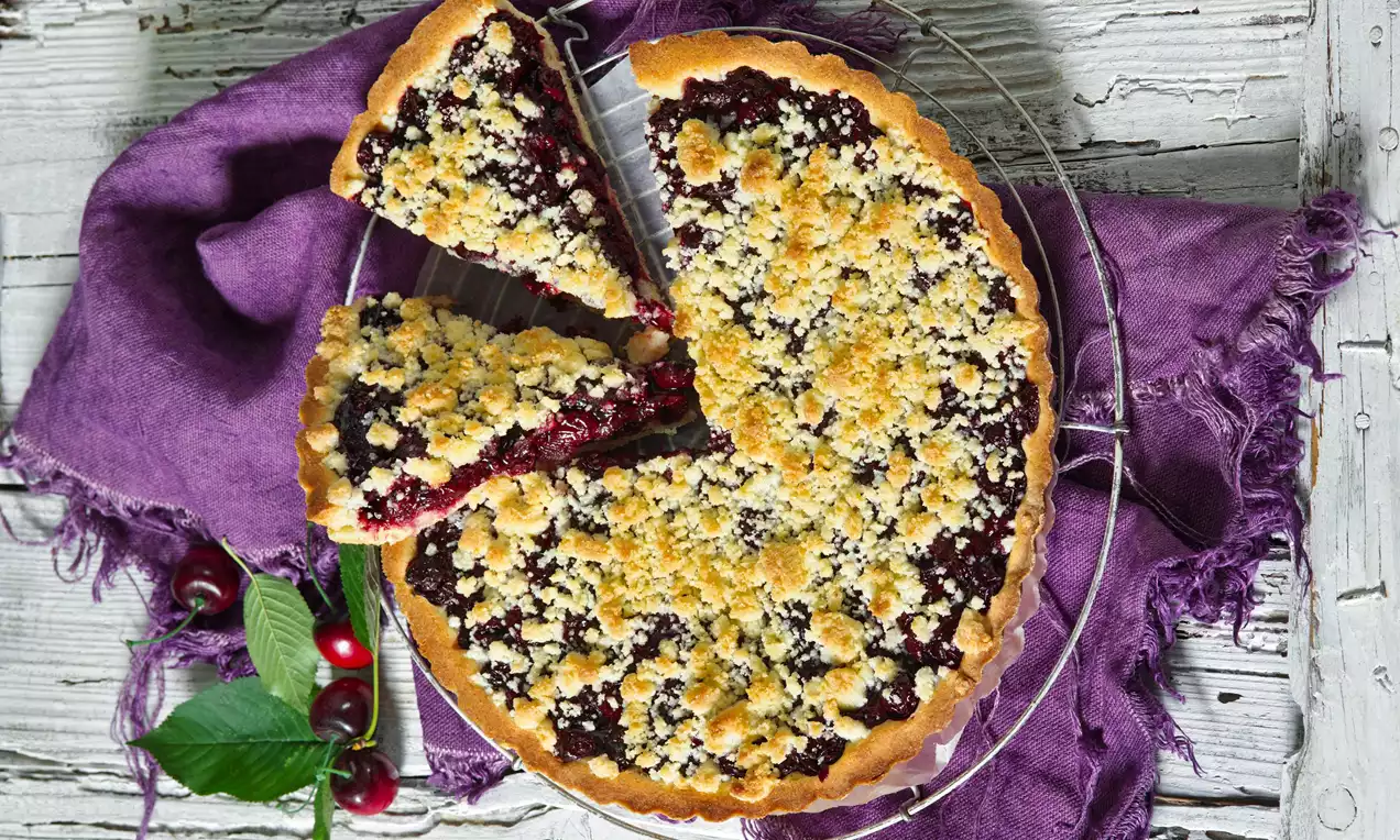 Šťavnatý višňový koláč na křupavém těstě s drobenkou, hluboká červená náplň, podávaný na drátěné mřížce s fialovou látkou a listy višní