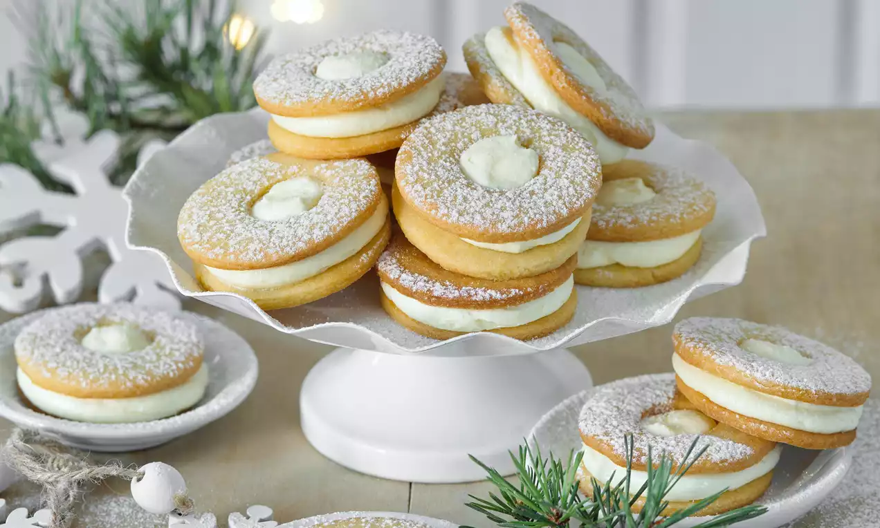 Kulaté sušenky světlé barvy s krémovou náplní uprostřed, posypané moučkovým cukrem na bílém stojanu, obklopené zimními dekoracemi