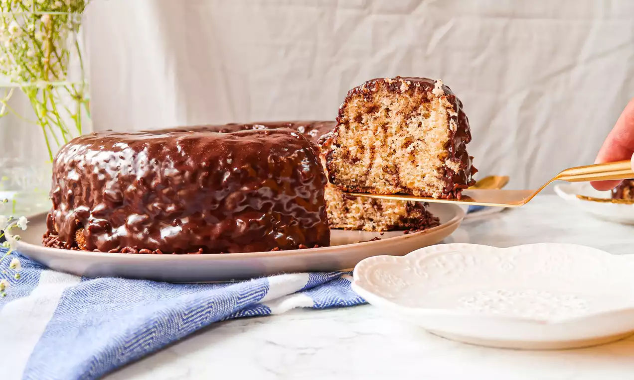 Bolo redondo com massa clara pontilhada de chocolate, coberto por calda espessa marrom brilhante, servido em prato branco sobre guardanapo azul