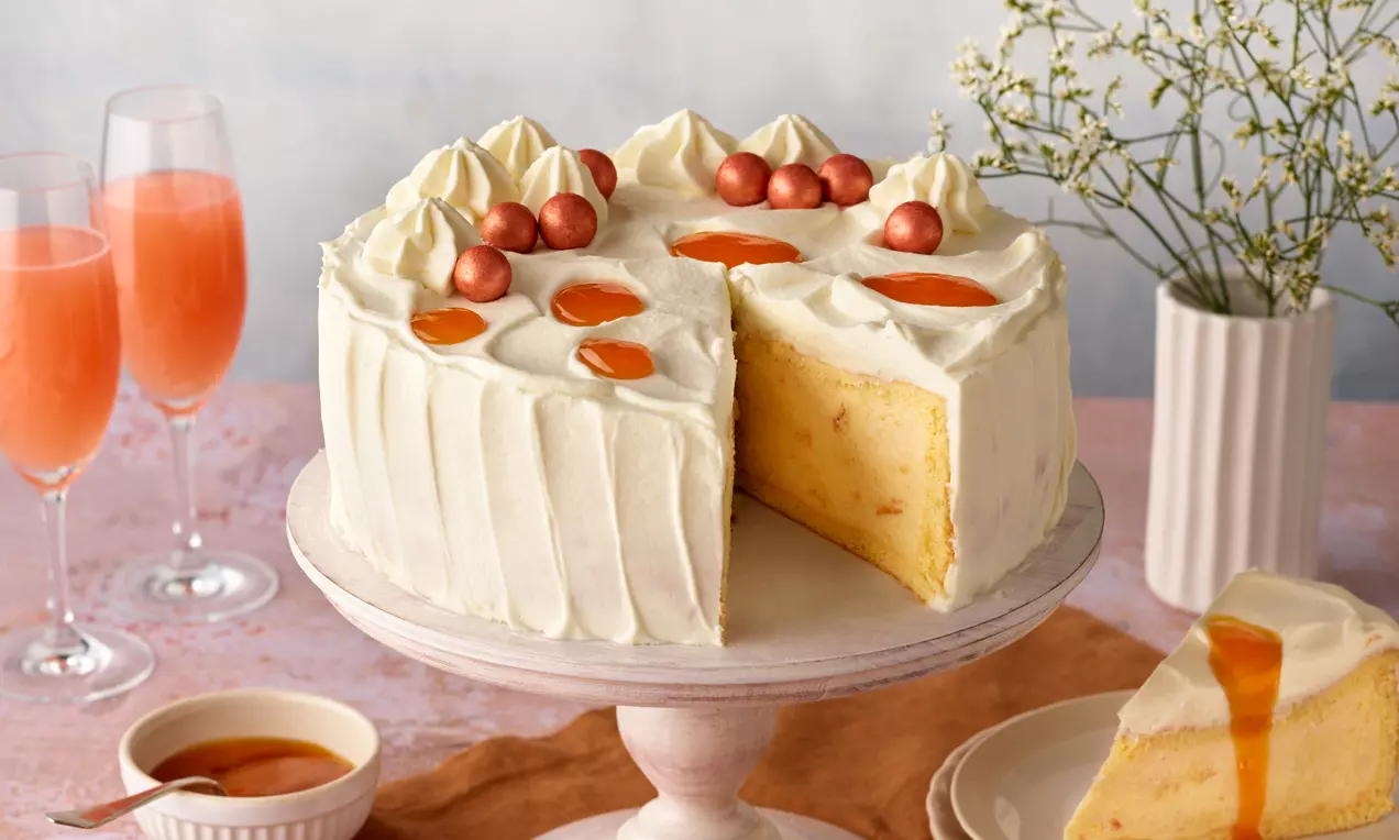 Bellini-Torte mit heller Creme, orangefarbenen Fruchttupfen und schimmernden Perlen auf weißer Tortenplatte, angeschnitten mit sichtbarer Füllung