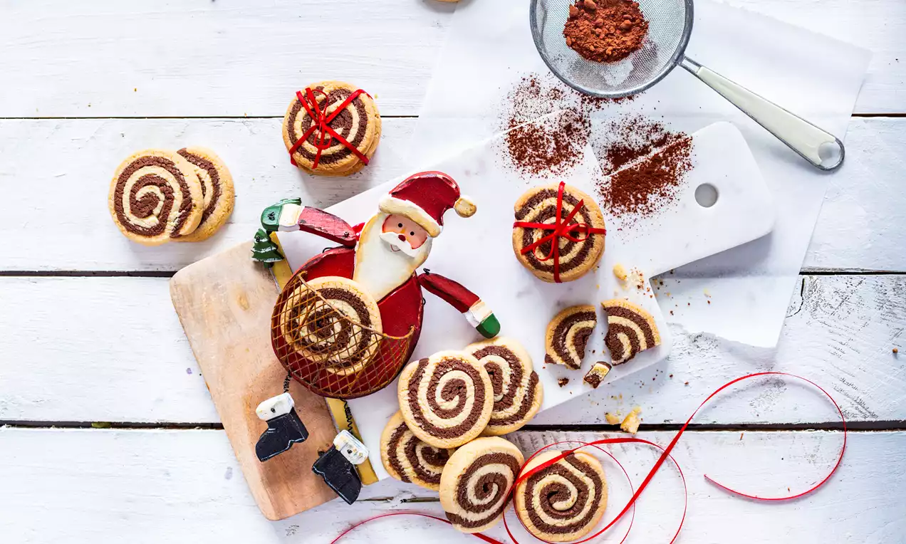 Veganski piškoti z dvojnim spiralnim vzorcem rjave in svetlo rumene barve, razporejeni na beli leseni podlagi ob figurici Božička