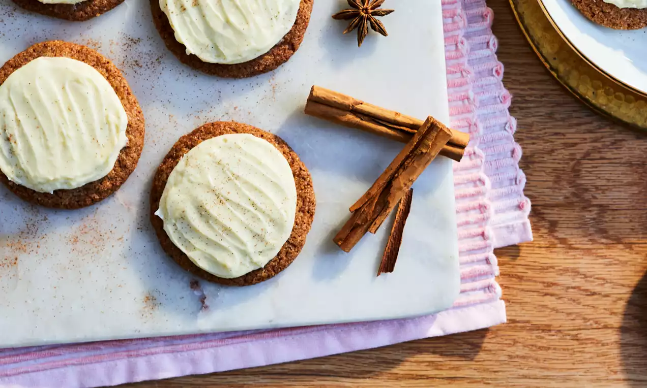 Chai cookies with spiced brown bases topped with creamy white frosting on a marble board surrounded by star anise and cinnamon sticks