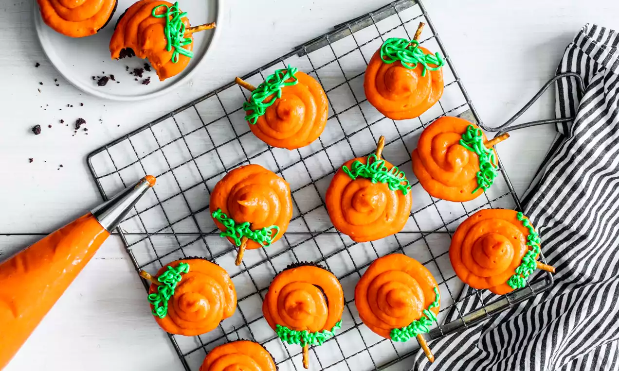 Helderoranje cupcakes met spiraalvormige glazuur, groene decoratie en pretzelstengels op een koele metalen rek naast een zwart-wit gestreepte doek
