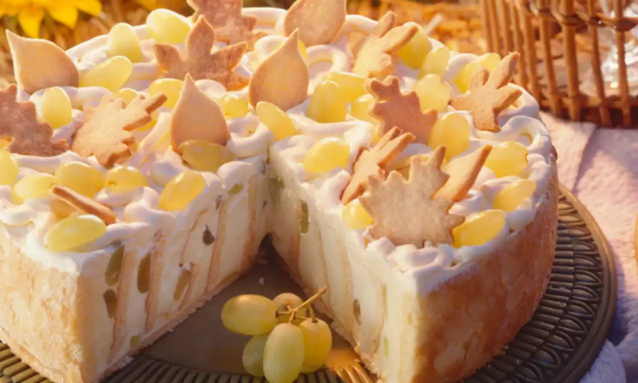 Gebäckrand mit heller Füllung, gelben Weintrauben und Plätzchen in Blattform, auf grünem Teller, umgeben von herbstlichem Dekor