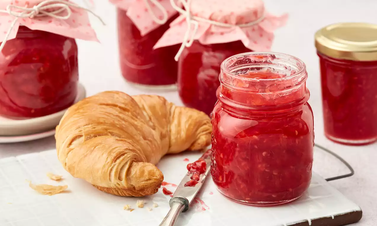 Bleščeča rdeča marmelada z drobnimi malinami v steklenem kozarcu ob zlatem, listnatem rogljičku in srebrnem nožu na beli deski