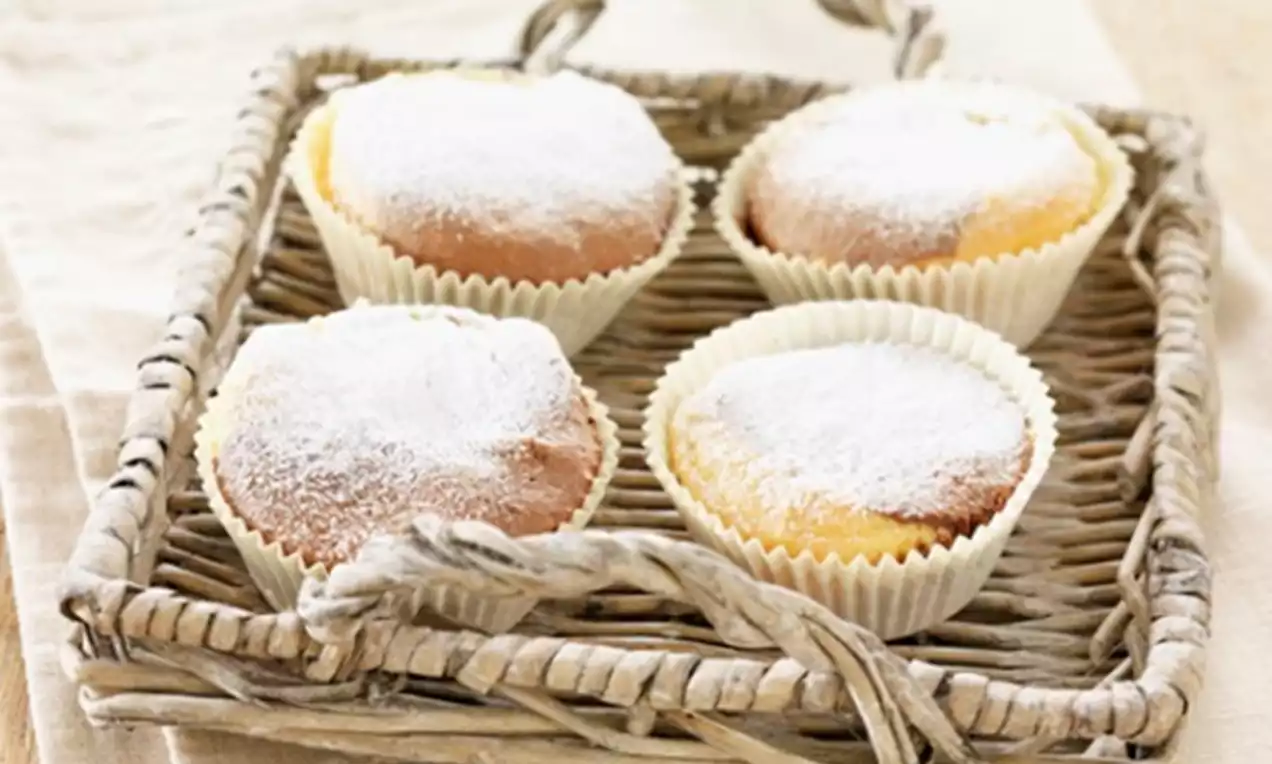 Luchtige choco-rumcakejes in witte bakpapiertjes, bestoven met poedersuiker, gepresenteerd in een rustiek gevlochten mandje op een licht tafelkleed