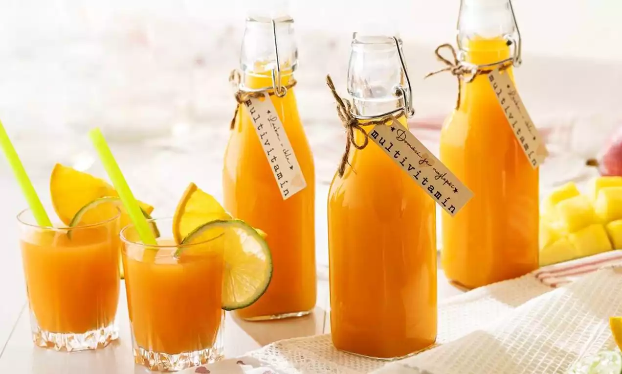 Lahve s jasně oranžovým sirupem s ručně psanými etiketami, skleničky s limetkovými plátky a brčkem, doplněné kostkami manga na bílém ubrusu