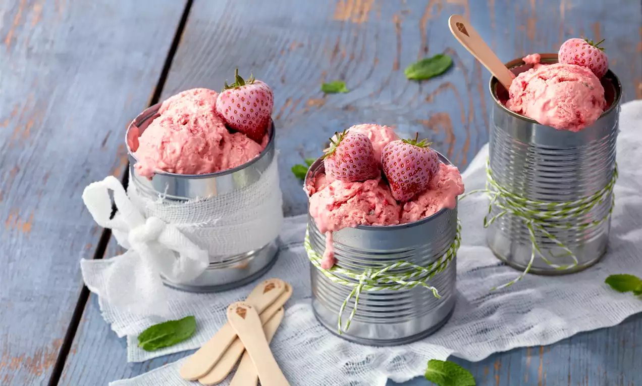 Erdbeer-Mascarpone-Eis in silbernen Dosen mit grüner Kordel, garniert mit frischen Erdbeeren auf weißem Tuch und Holztisch