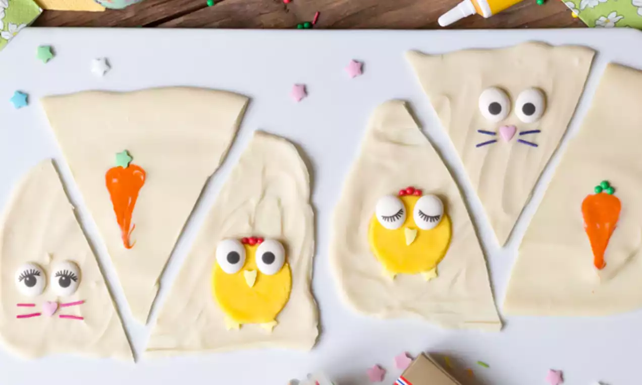 Vit choklad dekorerad som kycklingar och morötter med gula och orangea detaljer, placerad på en vit bricka omgiven av påskägg och dekorationer