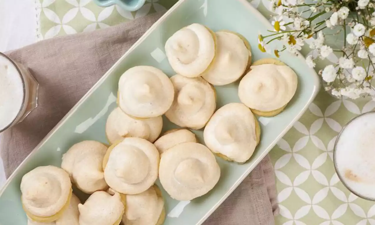 Hellgoldene Toffifee-Guetzli mit luftiger Baiserhaube auf einem mintgrünen Teller, umgeben von Blumen, Milchschaum und einer Stoffserviette