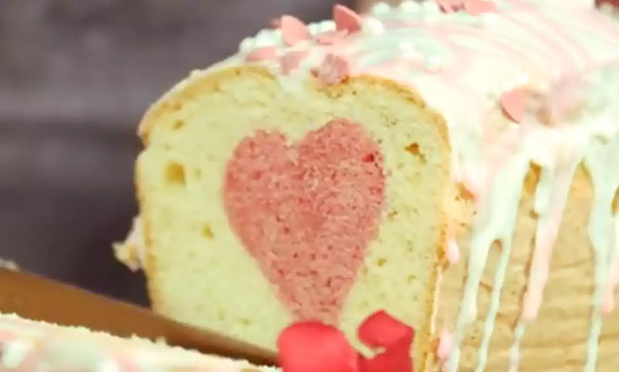 Kēkss gaiši zelta krāsā ar rožaini sarkanu sirds formu vidū, pārklāts ar baltu un rozā glazūru, rotāts ar cukura sirsniņām