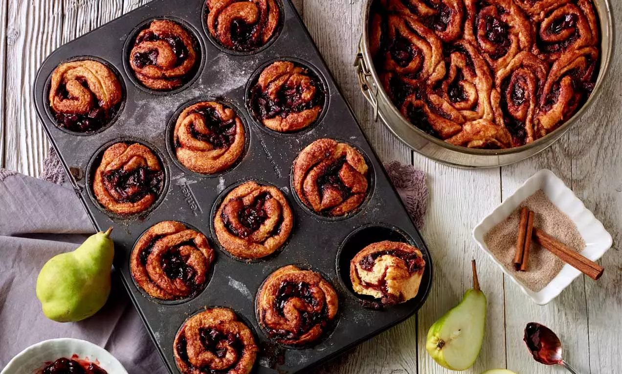 Kleine, goldbraune Schnecken mit dunkler Füllung in einer Muffinform und springform, dekoriert mit grünen Birnen und Gewürzen auf Holz