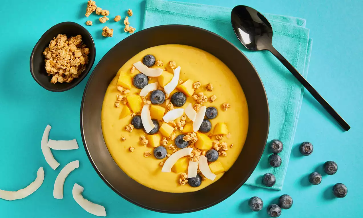 Protein-Smoothie-Bowl mit gelber Mango-Ananas-Creme, garniert mit Blaubeeren, Kokoschips und Granola in schwarzer Schale auf türkisem Hintergrund