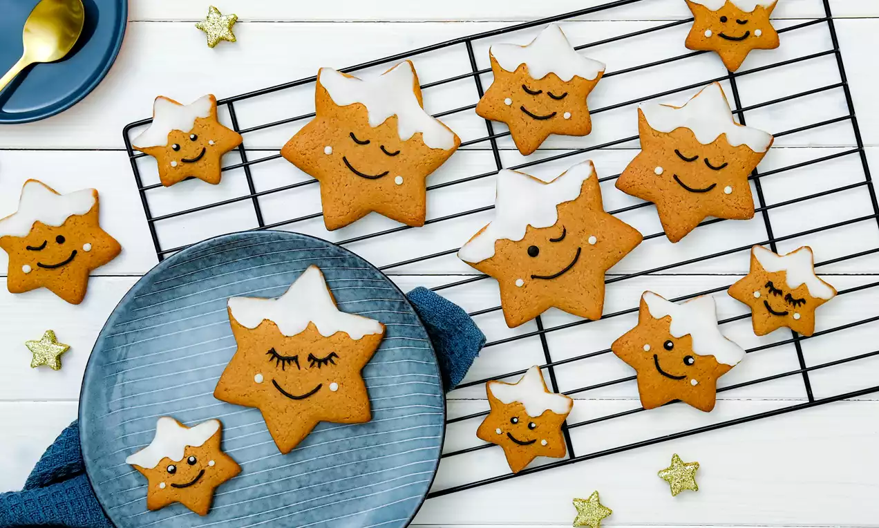 Biscuits étoilés brun doré avec glaçage blanc en forme de neige, décorés de visages souriants, disposés sur une grille et assiette bleue