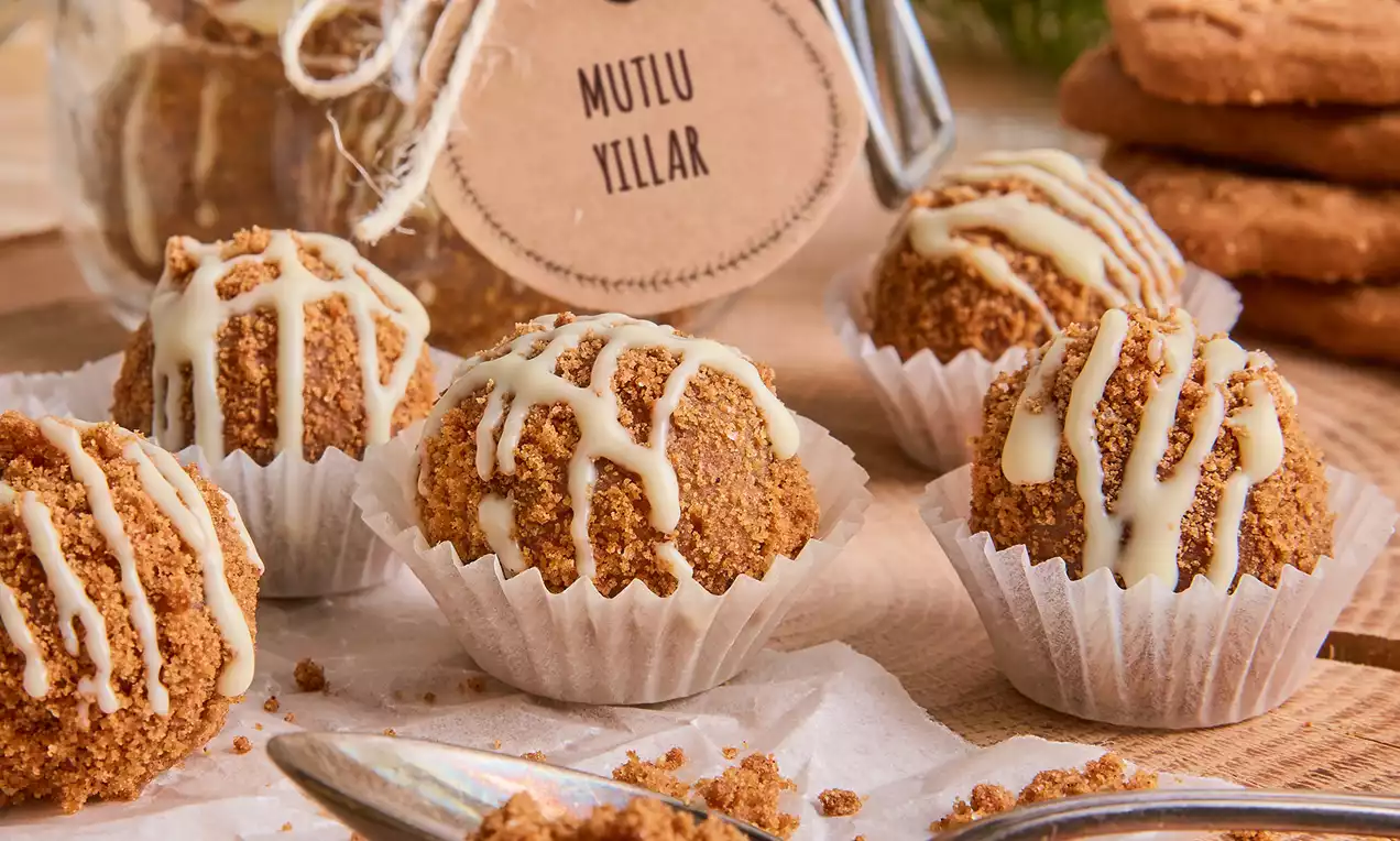 Beyaz kağıt kalıplarda sunulan, ince bisküvi kırıntılarına bulanmış, beyaz çikolata şeritleriyle süslenmiş yuvarlak bademli toplar