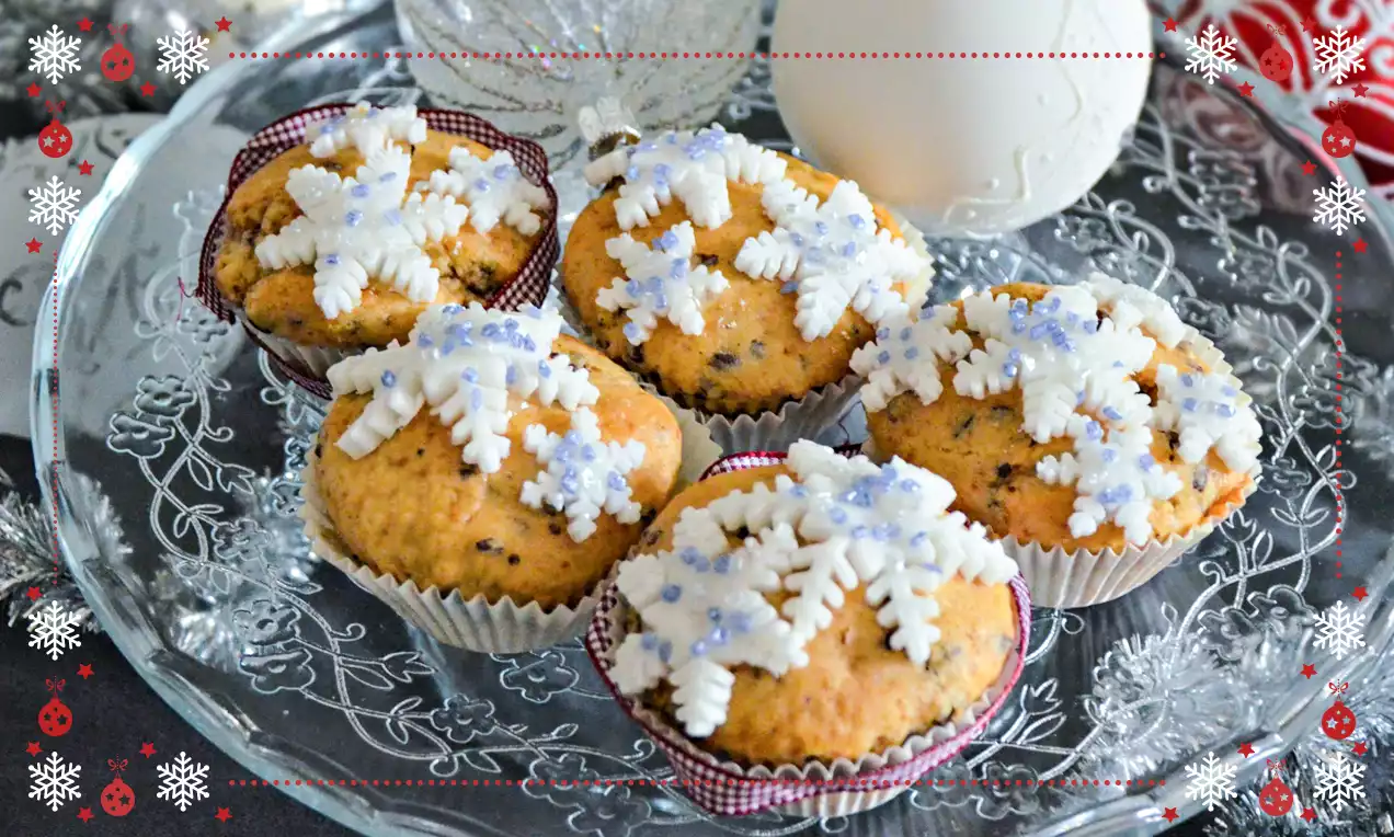 Muffin dorati guarniti con fiocchi di neve di zucchero bianchi e perline lilla, disposti su un vassoio di vetro decorato con motivi floreali
