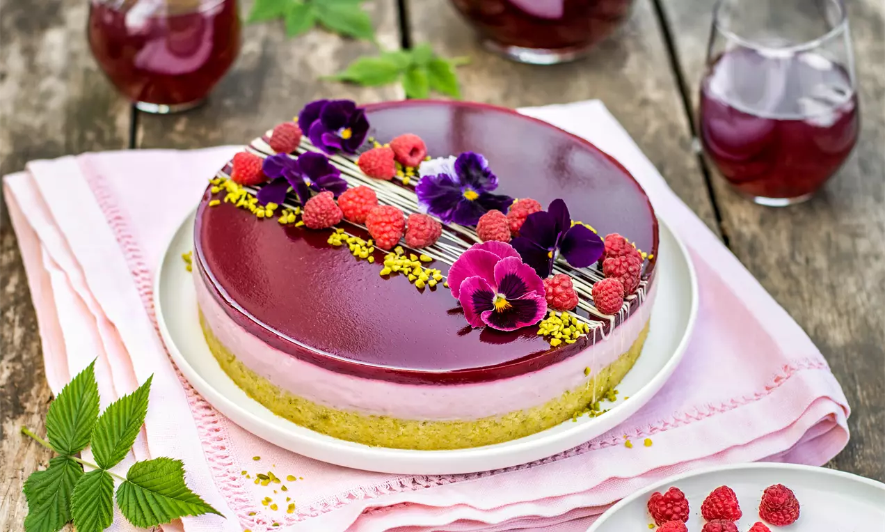 Малинов чийзкейк с лъскав рубиненочервен слой, кремообразно светлорозово ядро и бисквитен основа, украсен с свежи малини и виолетови цветя върху бяла чиния