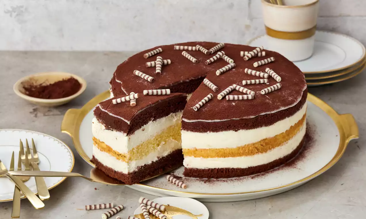 Dreischichtige Torte mit heller Creme, Schokoladenboden und Kakaopulver, dekoriert mit weißen Schokoröllchen auf runder Servierplatte