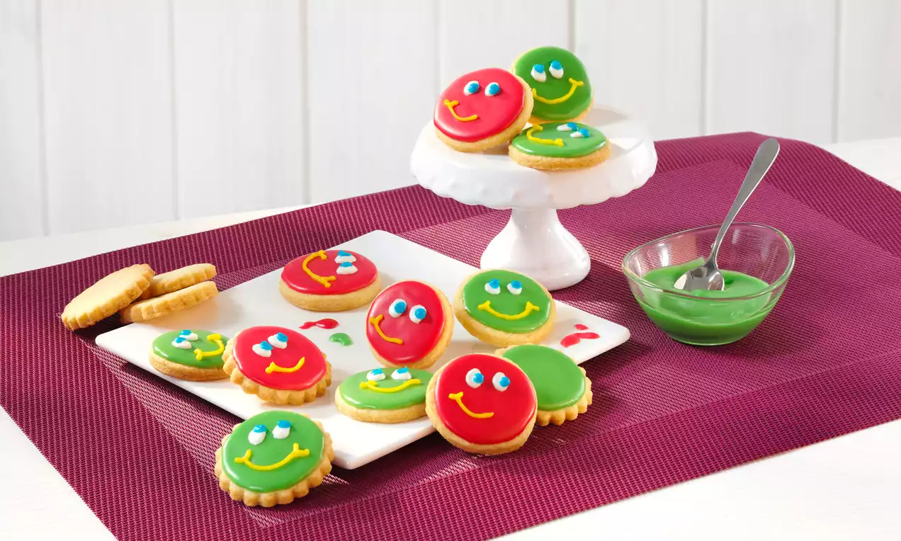 Runde Mürbeteigkekse mit glatter roter und grüner Glasur, lächelnden Gesichtern und Zuckeraugen auf einem Teller und einer weißen Etagere