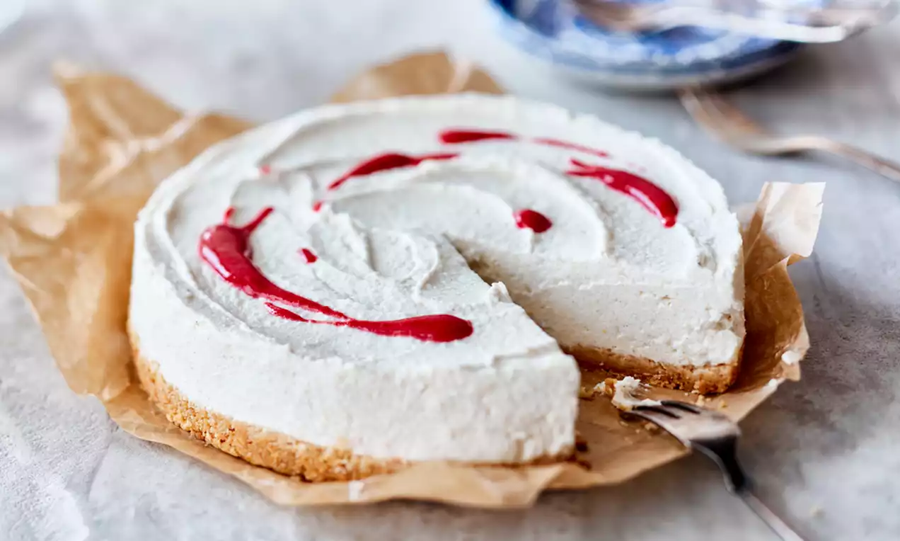 Cheesecake de vanilie cu blat crocant auriu, cremă albă fină și dungi ondulate de sos roșu pe hârtie pergament, lângă farfurie decorativă albastră