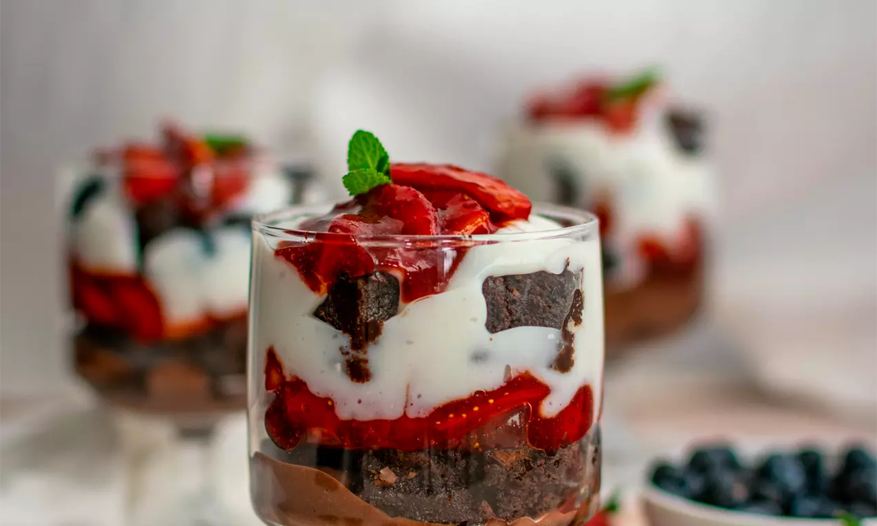 Брауни трайфъл в стъклена чаша със слоеве тъмен брауни, бял лек крем, яркочервени намачкани ягоди и свежа мента отгоре, заобиколен от плодове