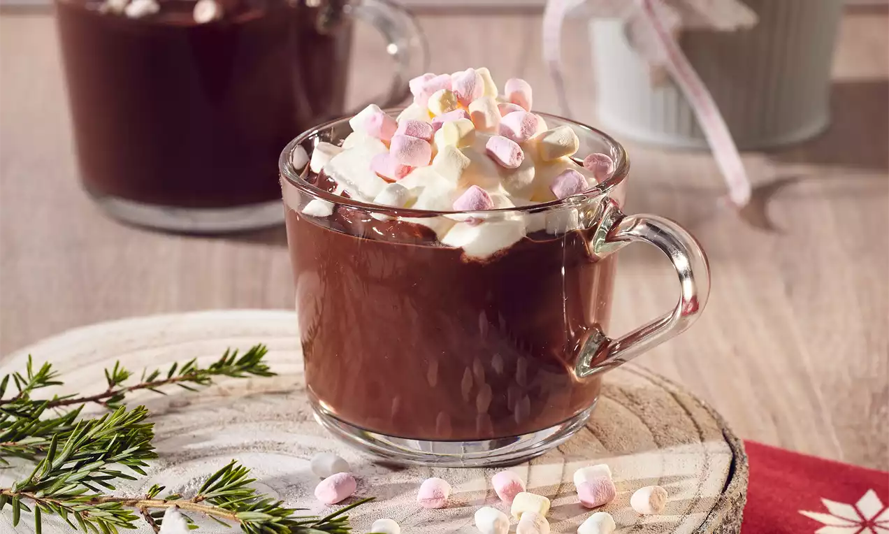 Skleněný hrnek plný tmavě hnědého kakaa, ozdobený vrstvou bílé šlehačky s růžovými a bílými mini marshmallows na dřevěném podnosu