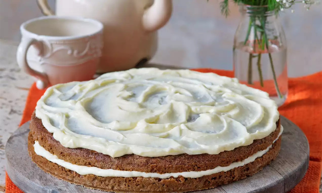 Mrkvový koláč s jemným smotanovým krémom vo vlnitých vrstvách na drevenom podnose s oranžovým obrúskom, v pozadí čajník a kvetiny
